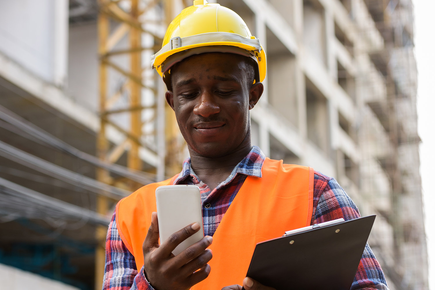 Young black African man construction worker holding clipboard while using mobile phone at building site